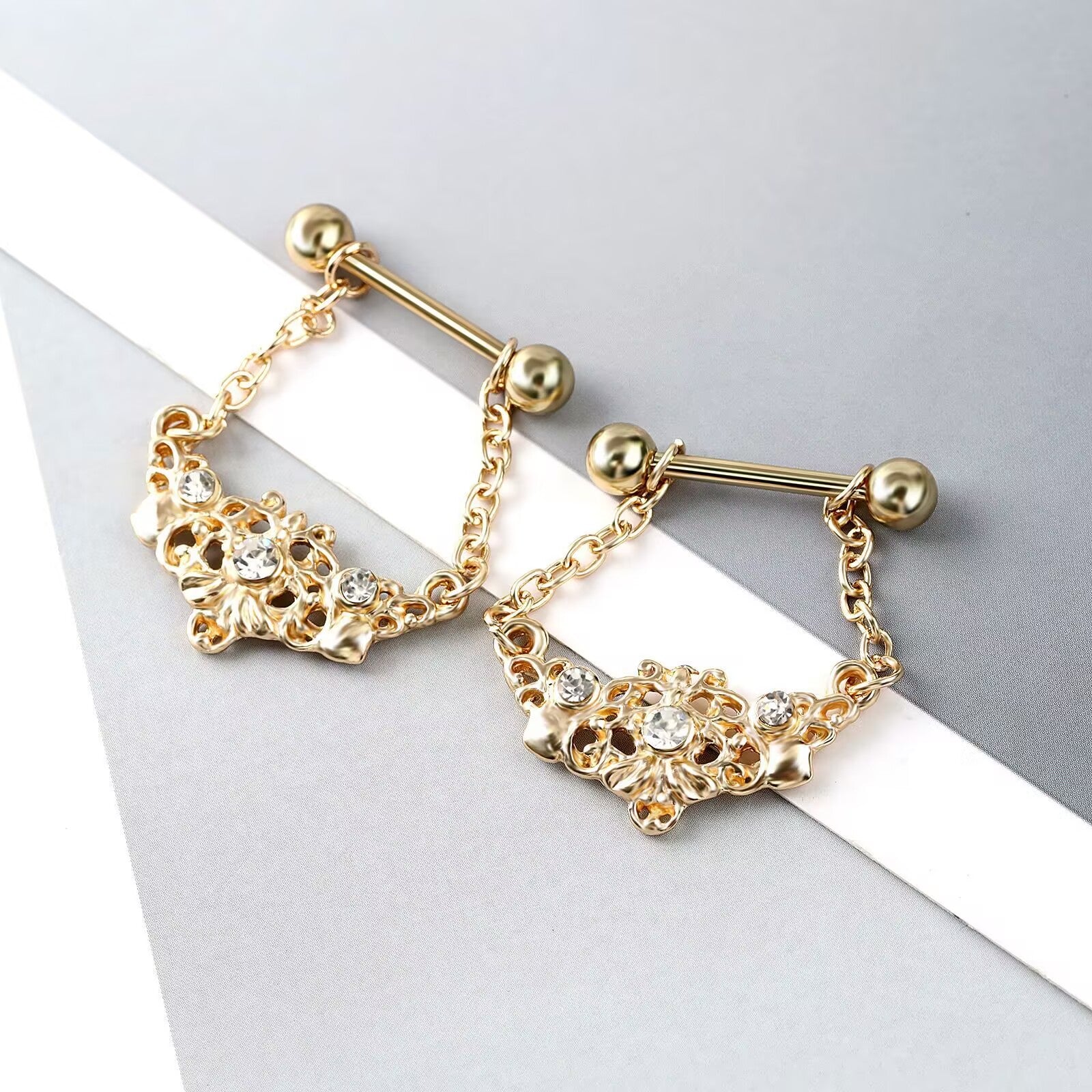 Close-up of gold gothic nipple barbells resting on a grey and white aesthetic background