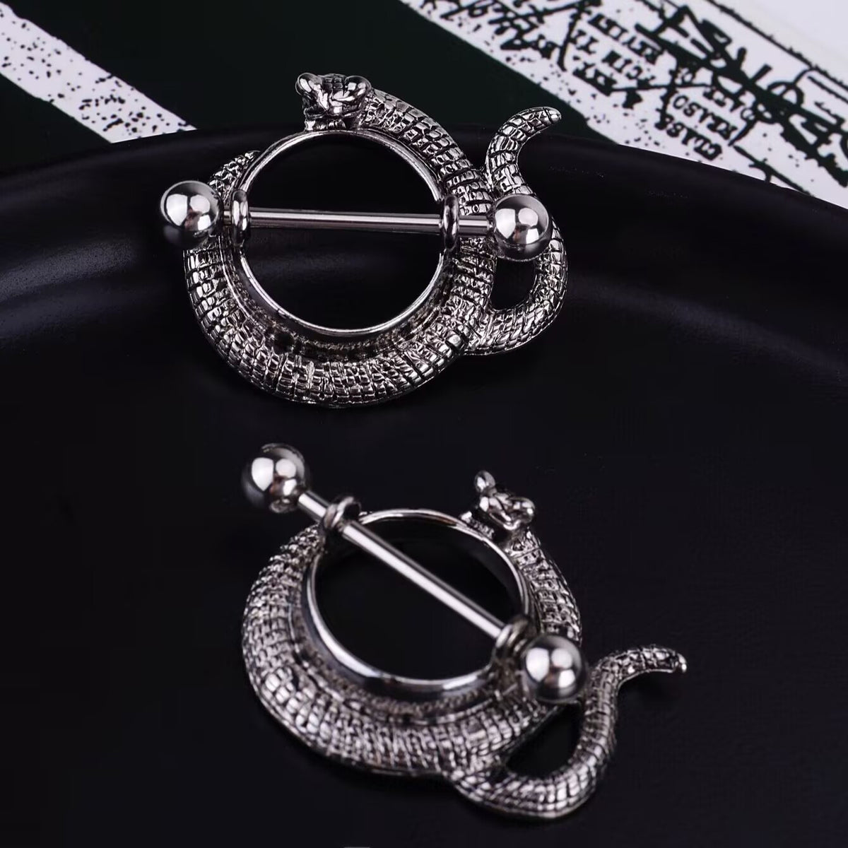Two snake-shaped nipple shields on a black tray with a gothic script background