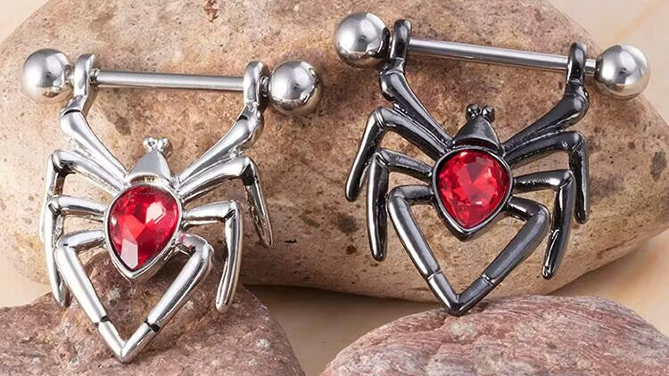 Three different spider nipple shield rings (silver, black, rainbow) arranged on decorative stones.