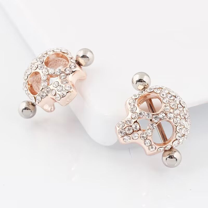 A pair of rose-gold skull nipple shields with crystal pave, displayed on a white surface at slightly different angles.
