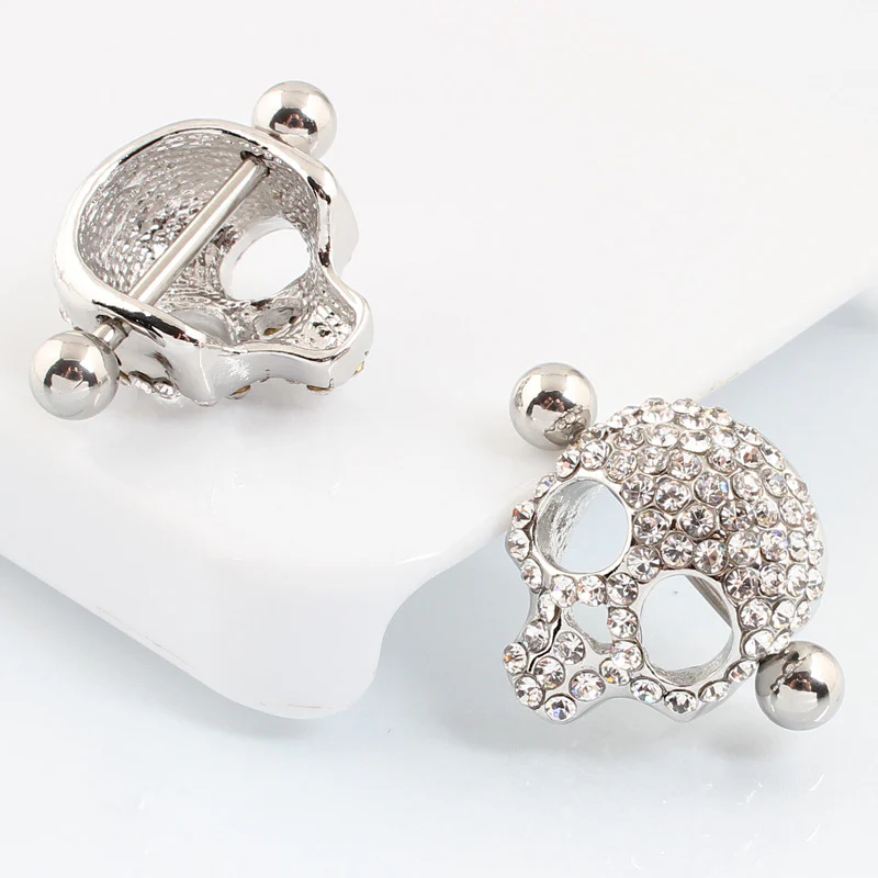 A detailed view of a pair of silver crystal skull nipple shields, with one positioned to show the back construction and barbell, and the other front-profile.