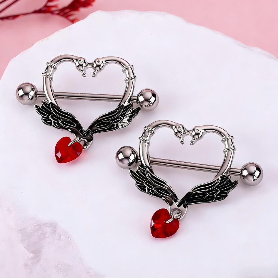 Pair of silver swan heart nipple shields with black wings and red heart charms on a white stone background.